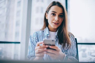 Modern cep telefonu cihazını elinde tutan ve e-posta bültenini beklerken kameraya bakan genç, güzel bir kadın blogcunun yarım boy portresi.