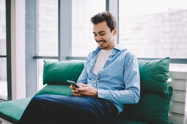 Rahat koltukta dinlenen cep telefonuyla günlük kıyafetlerle e-posta yazan neşeli hippi adam online platformda bilgi arayan başarılı erkek blogcu.