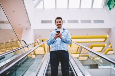 Modern yürüyen merdivenden inerken kameraya gülümseyen neşeli beyaz adamın yarım boy portresi alışveriş öncesi kredi kartındaki bakiyeyi kontrol etmek için akıllı telefon tutuyor.