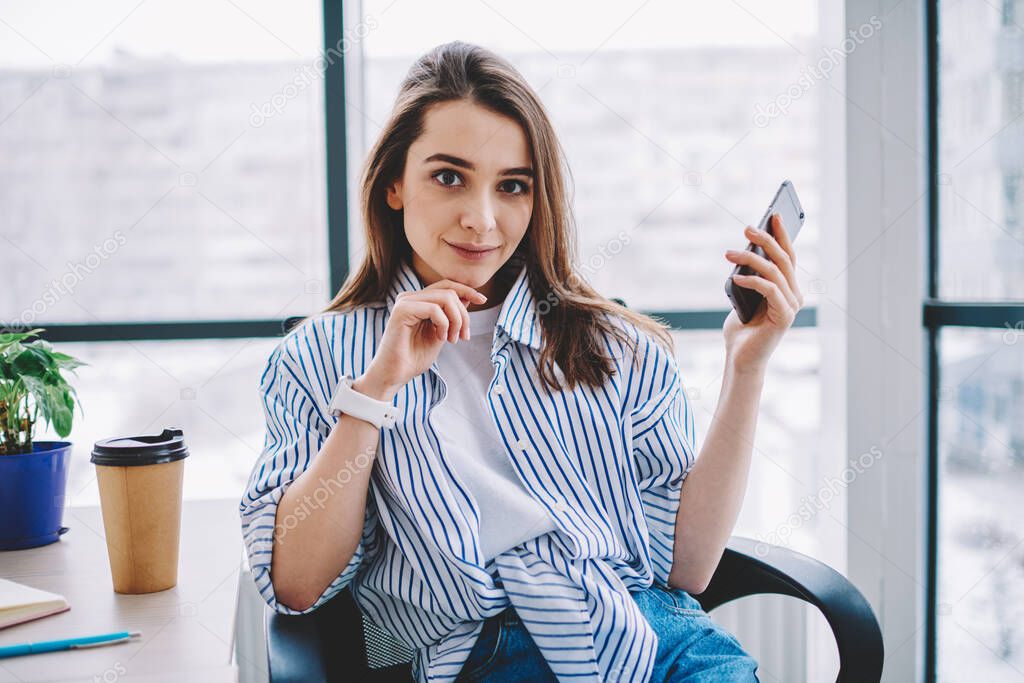 Portrait of Caucasian female employee in smart casual outfit sitting at table desktop with disposable takeaway cup and looking at camera during job break for social networking, wireless mobility