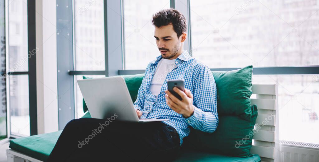 Caucasian hipster guy sharing information via cellular phone and laptop device using bluetooth connection while working remotely indoors, skilled male reading incoming message via app on netbook