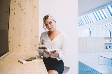 Modern ofis sunum odasındaki ahşap tezgahta otururken iş kataloglarından bilgi okuyan kendinden emin genç profesyonel kadın yönetmen.