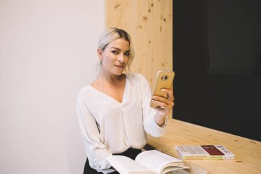 Çalışma alanında poz verirken modern cep telefonu cihazları ve edebiyat kitabı tutan güzel kadın kullanıcının portresi, akıllı telefon teknolojisiyle kameraya bakan akıllı okuyucu