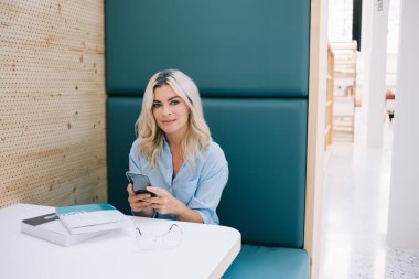 Üniversite sınavlarına hazırlanan zeki kız öğrencilerin portresi kütüphanede edebiyat kitabı ve akıllı telefonla bilgi araştırması, öğrenim ve öğrenim yapmak için masada oturuyor.