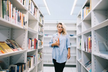 Gözlüklü, kendine güveni tam genç bir bayan kitap tutuyor ve kütüphanede beyaz kitaplıklar arasında dururken kameraya bakıyor.