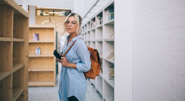 Sırt çantası ve edebiyat kitaplıklarının yanında en çok satanlar arasında poz veren modaya uygun giyinmiş kadın portresi klasik gözlüklü kız öğrenci akıllı telefon cihazı tutuyor.