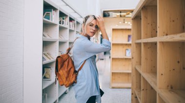 Görsel koruma için optik gözlüklü akıllı kadın okuyucunun yarı boy portresi halka açık kitapçıda alışveriş yaparken kameraya bakıyor, sırt çantalı güzel bir kadın içeride poz veriyor