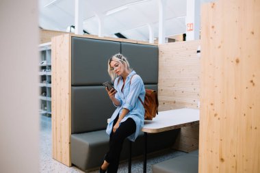 Barcelona 'daki modern ışık kütüphanesi odasındaki masaya yaslanırken cep telefonundan mesaj atan günlük kıyafetlerle konsantre genç bayan öğrenci.