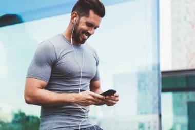 Elektronik kulaklıklı neşeli beyaz vücut geliştirici akıllı telefon uygulaması aracılığıyla olumlu dostça sohbet sırasında sesli mesaj aldı, mutlu erkek atlet 30 'lu yaşlarda cep telefonu mesajları gönderiyor.
