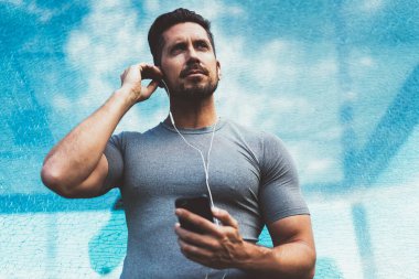 Kafkasyalı spor blogcusu elinde akıllı telefon cihazıyla dikkatle bakıyor sabah sporuna hazırlanıyor elektronik kulaklıklı kaslı atletik adam mobil motivasyon podcast 'ini dinliyor.