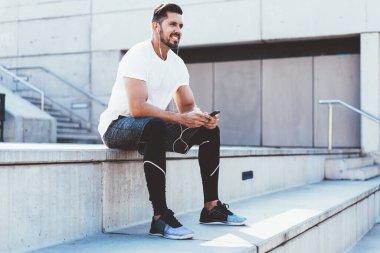 Mutlu sakallı sporcu, müzik listesindeki şarkıları seviyor. Kulaklıklara bağlı, eğitimden sonra dinleniyor. Müzik setleri ve cep telefonu uygulamalarıyla online radyo dinleyen gülümseyen bir erkek koşucu.