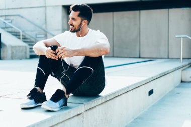 Mutlu sakallı sporcu, müzik listesindeki şarkıları seviyor. Kulaklıklara bağlı, eğitimden sonra dinleniyor. Müzik setleri ve cep telefonu uygulamalarıyla online radyo dinleyen gülümseyen bir erkek koşucu.