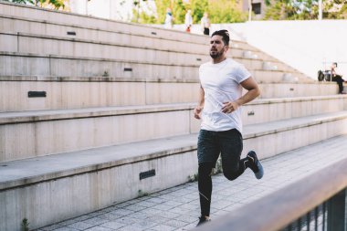 Aktif giyimli beyaz erkek şehir caddesinde koşuyor, kulaklıklı sporcu müzik listesi şarkıları dinliyor sağlıklı yaşam tarzını ve mükemmel vücut şeklini korumak için kardiyo eğitimi alıyor.