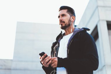 Elektronik kulaklıklar takan, şehir caddesinde durup, canlılık egzersizini düşünen, 30 yaşında, beyaz vücutçu, spor sesli kitap dinleyen, tefekküre yatkın deneyimli bir antrenör.