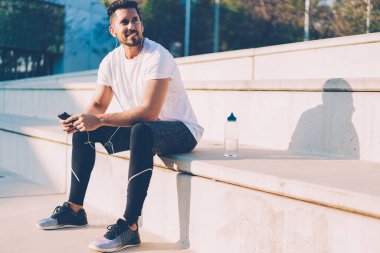 Neşeli bir beyaz atlet elinde hücreyle güneşli bir şehirde dinleniyor ve gülümsüyor. Elektronik kulaklıklı kaslı bir erkek vücut geliştirici. Açık havada müzik dinlemenin keyfini çıkarıyor.