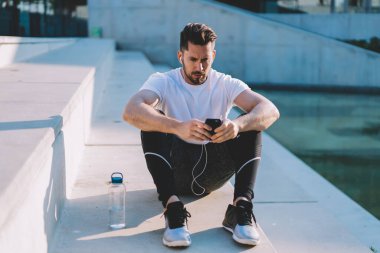 Sağlıklı yaşam tarzı hakkında motivasyon videosu izlemek için elektronik kulaklık kullanan kaslı vücut çalıştırıcı, akıllı telefon web sitesinde spor haberlerini kontrol eden 30 'lu yaşlarda kulaklık takan beyaz vücutlu vücut geliştirici.