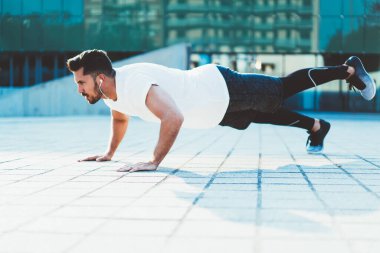 Kaslı vücutlu güçlü beyaz sporcu sokaklarda şınav çekiyor. Sabah antrenmanının keyfini çıkarıyor. Elektronik kulaklıklı güçlü erkek sporcu, canlılığını ve sağlığını etkinlikte tutuyor.