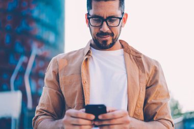 Görme düzeltmesi için gözlüklü modacı şehir merkezinde cep telefonunda uygulamayı güncelliyor ve internet bağlantısı kullanıyor, beyaz erkek blogcu web sohbeti sırasında mesaj yazıyor