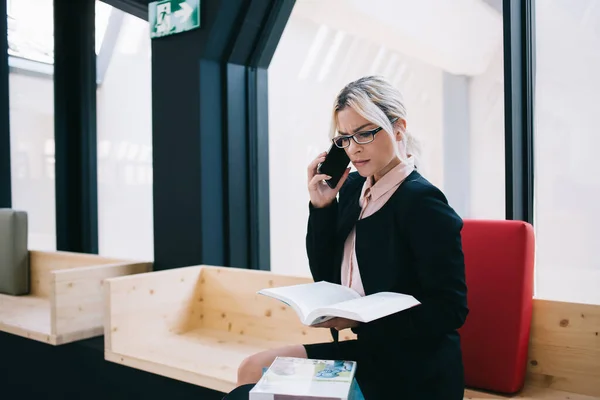 Puzzled female secretary dressed in formal wear calling to corporate boss for verify organisation plan from textbook, woman in eyewear using informative notepad for discussing business while phoning