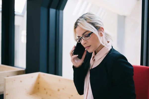 穿着黑色西服和眼镜的严肃时髦的女企业家坐在现代工作空间的窗边打电话 — 图库照片