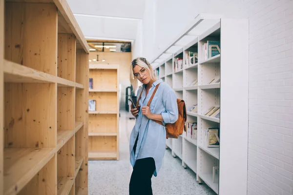 图书馆里穿着休闲装 背着背包 头戴眼镜 站在书架旁边 望着相机的时髦女性侧视图 — 图库照片