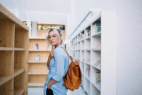 Half length portrait of intelligent female reader in optical eyewear for vision protection looking at camera during time for shopping in public bookstore, beautiful woman with backpack posing indoors