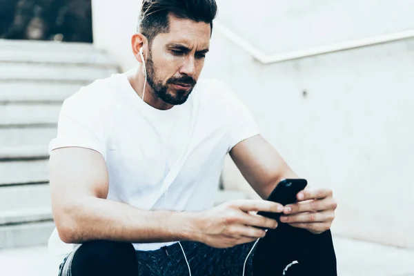 Caucasian male jogger in tech earphones connecting to 4g wireless for consultancy messaging with personal online trainer, masculine man with cellular installing app for listening sportive audio guide