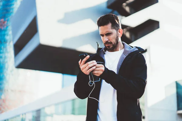 Male athlete in active wear checking notifications on mobile phone having break after workout outdoors, sportsman in earphones sending mail message while download media music to smartphone player