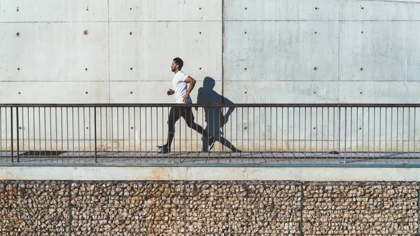 在城市晨练时穿着运动服跑步的精力充沛的男性运动员 在街上慢跑时拿着耳机听音乐的有动力的运动员的侧视图 — 图库照片