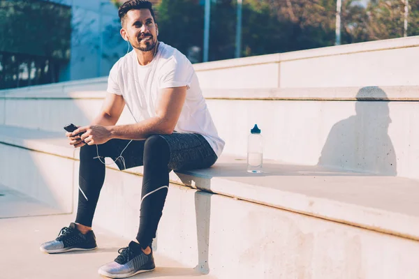 Neşeli bir beyaz atlet elinde hücreyle güneşli bir şehirde dinleniyor ve gülümsüyor. Elektronik kulaklıklı kaslı bir erkek vücut geliştirici. Açık havada müzik dinlemenin keyfini çıkarıyor.