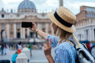 Sırt çantalı, çizgili tişörtlü ve şapkalı mutlu genç bayan turistin Vatikan 'da kare üzerinde zafer jesti ve akıllı telefonuyla selfie çekerken yan görüntüsü.