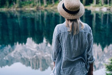 Şapkalı ve çizgili tişörtlü genç bir kadının, güneşli bir günde, Karersee Gölü kıyısında güneşli bir dağ sırtının yansımasıyla duruşu.