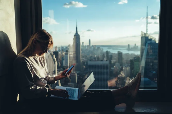 Young distance worker sharing multimedia files via mobile and laptop technology connecting to bluetooth, Caucasian freelancer messaging on smartphone sitting at panoramic window in New York