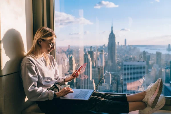 Young distance worker sharing multimedia files via mobile and laptop technology connecting to bluetooth, Caucasian freelancer messaging on smartphone sitting at panoramic window in New York