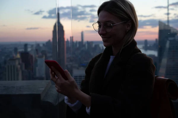 ニューヨークを訪問するためのソロ旅行の旅の間にローミングインターネットに接続するフォロワーとのソーシャルメッセージのためのセルラーデバイスを使用して笑顔の女性観光客 陽気なヒップスター女の子のブラウジングウェブ — ストック写真