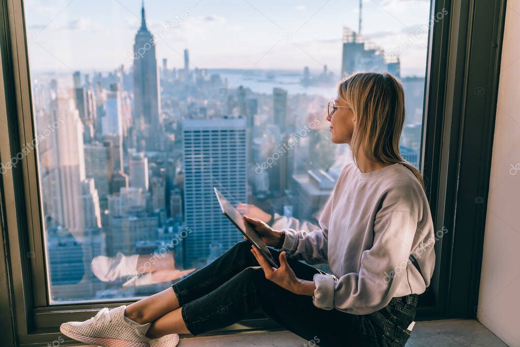Pondering millennial blogger holding modern digital tablet and dreaming during travel journey in New York looking on Manhattan district from above, youthful woman with technology thinking indoors