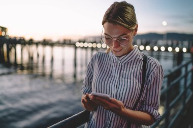 Gün batımında rıhtımda durup tatil boyunca dinlenirken akıllı telefonları karıştıran günlük kıyafetlerle ve gözlüklerle gülümseyen bir kadın.