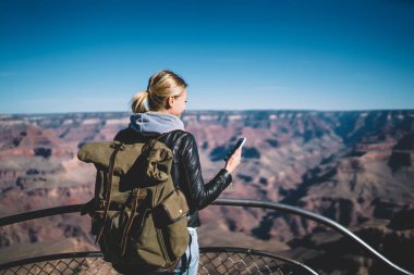 Büyük sırt çantalı, deri ceketli sarışın kadın gezgin Grand Canyon Ulusal Parkı 'nda duruyor ve akıllı telefonları tarıyor.