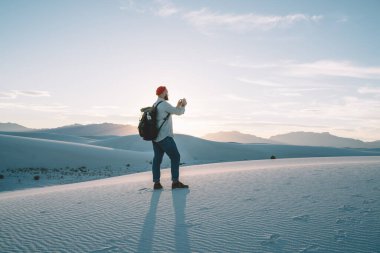 Beyaz Kumlar Ulusal Parkı 'nda yanan dağların arkasında dikilirken çevresindeki manzaranın fotoğraflarını çeken kırmızı şapkalı yüzü olmayan sakallı adamın arka plan görüntüsü.