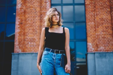 Düşük açılı, odaklanmış, gözlük takan, elinde not defteriyle tuğla bina karşısında bekleyen ve gözünü kaçıran genç bir kadın.