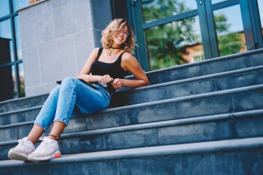 Sıradan kıyafetli, gözlüklü, gülümseyen ve üniversitenin merdivenlerine yaslanarak eğlenen genç bir bayan.