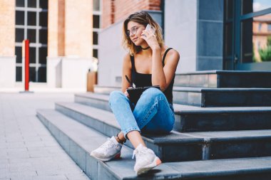 Rahat giysiler içinde düşünceli, narin bir genç kız. Fotokopi defterine not alıyor ve merdivenlerde bacak bacak bacak üstüne atarak otururken telefonla konuşuyor.