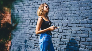 Gündelik giysiler ve güneş gözlükleriyle lolipop takmış, akıllı telefondan kulaklıkla müzik dinleyen ve parlak güneşin altında serinleyen çekici genç bayan milenyumu.