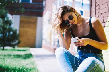 Esmer kadın kulaklıklı, akıllı telefon kullanan, saçları düzleştiren ve yazın çimenlikteki evin yanında dinlenen.