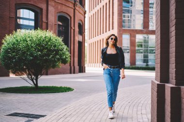 Güneş gözlüklü, sıradan giysileri olan ve modern şehrin caddeleri boyunca binaların arasında yürüyen genç bir kadın vücudu.