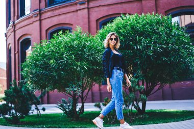 Güneşli bir günde evin yakınındaki çimenlikte yeşil çalıların yanından geçen gündelik giysili esmer kadın yan görünüşü.