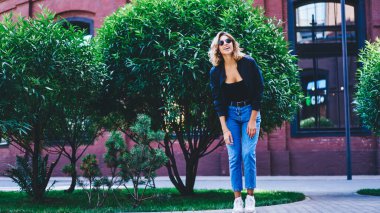 Modaya uygun giyinmiş, gülümseyen ve caddede bina ve çalılara karşı duran siyah güneş gözlüklü genç bir kadın vücudu.