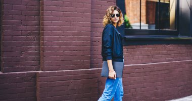 Gündelik kıyafetleri ve güneş gözlükleriyle modern binanın yanında elinde not defteriyle dikilip kameraya bakan modaya uygun genç bir kadın.