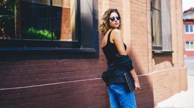 Moda kıyafetleri ve güneş gözlükleriyle sokakta dikilip kameraya bakarken deri ceketli genç bir kadının yan görüntüsü.