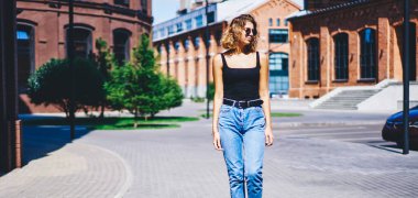 Düşünceli, genç, sıska, gündelik giysiler ve güneş gözlüğü takmış, güneşli bir günde kaldırımda yürüyen ve gözlerini çeviren bir kadın.